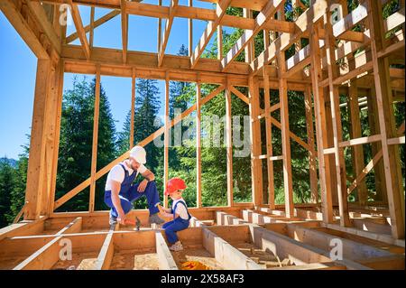 Père avec son fils en bas âge construisant une maison à ossature en bois. Constructeur masculin démontrant à son fils comment utiliser un marteau pour pilonner des clous sur le chantier de construction, portant des casques et des salopettes bleues le jour ensoleillé. Banque D'Images