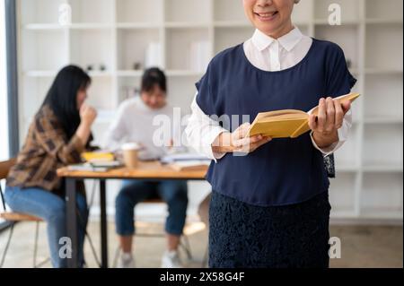 Une image recadrée d'une professeure asiatique souriante et satisfaite debout dans la salle de classe avec un livre à la main pendant que les étudiants étudient. Banque D'Images