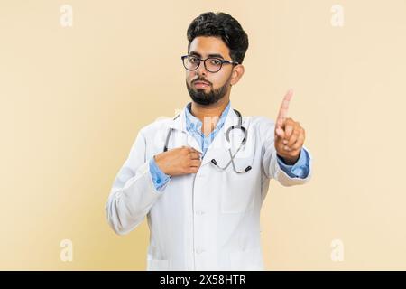 Je ne le veux pas, pas moi. Confus jeune médecin indien cardiologue homme pointant les doigts demander qui pourquoi moi non merci je n'ai pas besoin de refus signe d'arrêt. Apothicaire Guy sur fond beige Banque D'Images