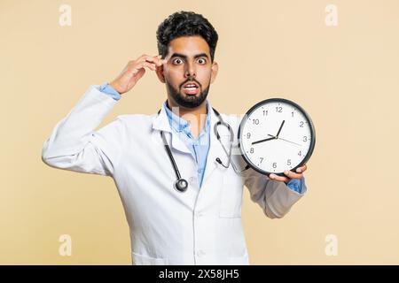 Jeune médecin indien cardiologue homme avec l'anxiété vérifiant le temps sur l'horloge, courant en retard au travail, étant en retard, délai. Mec de pharmacie arabe inquiétant d'être ponctuel isolé sur fond beige Banque D'Images