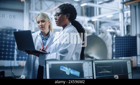 Deux femmes ingénieurs parlent, utilisent l'ordinateur travaillant à l'usine de fabrication de satellites aérospatiaux. Équipe diversifiée de scientifiques multiethniques effectuant des recherches scientifiques et technologiques pour le programme d'exploration spatiale Banque D'Images