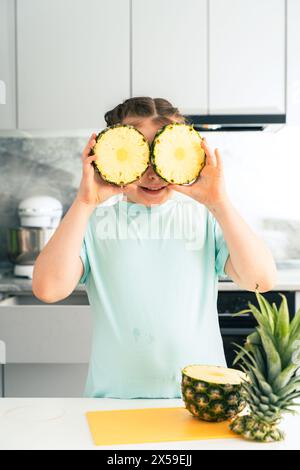 Une petite fille couvre ses yeux de cercles d'ananas. Une jeune fille pré-adolescente joue avec un ananas dans la cuisine à la maison. Foyer sélectionné. Phot de haute qualité Banque D'Images