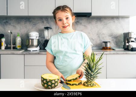 Heureuse petite fille coupant l'ananas sur le fond de la cuisine à la maison. Fille pré-adolescente souriante tout en préparant des fruits sains pour manger. Sélectionner Banque D'Images