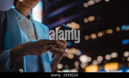 Concentrez-vous sur la main féminine à l'aide d'un smartphone marchant dans Night City Street pleine de néon Light. Femme tenant téléphone portable, affichage de médias sociaux, achats en ligne, SMS. Banque D'Images