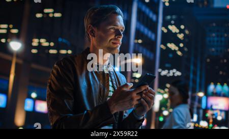 Portrait de beau jeune homme à l'aide d'un smartphone debout dans la rue de la ville de nuit pleine de néons lumières. Sourire élégant Blonde Male à l'aide du téléphone portable pour la publication sur les médias sociaux. Coup d'angle néerlandais. Banque D'Images