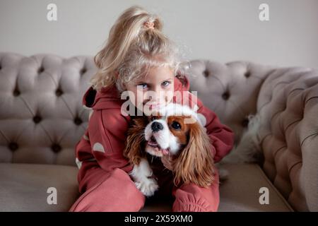 Belle fille blonde enfant est assis à la maison sur un canapé, jouant avec et embrassant le chien, créant des moments de joie et de compagnie avec l'animal de compagnie. Banque D'Images