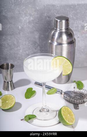 Cocktail d'alcool aux agrumes Gimlet. Boisson alcoolisée sucrée et aigre, avec gin ou vodka et garniture de citron vert, sur gris clair avec lumière dure et utensi de bar Banque D'Images