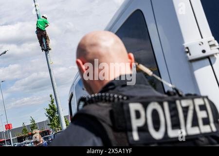 08 mai 2024, Brandebourg, Grünheide : un policier bloque l'accès à une action des militants de l'organisation de protection de la nature Robin Wood avec une bannière avec le texte 'Verkehrswende statt Autokonzerne! Robin Wood' comme une action de protestation contre l'expansion de l'usine Tesla à Grünheide à travers un parking devant les locaux de Tesla. Cette manifestation fait partie d'une semaine de manifestations contre Tesla. Photo : Carsten Koall/dpa Banque D'Images