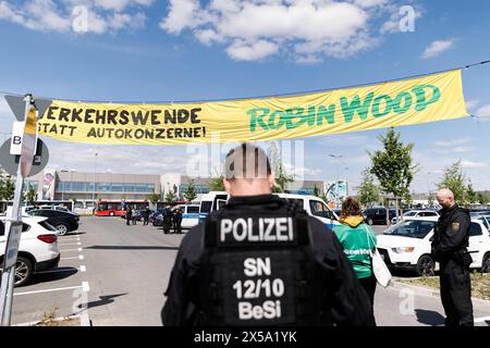 08 mai 2024, Brandebourg, Grünheide : des policiers sécurisent une action des militants de l'organisation de conservation de la nature Robin Wood avec une bannière avec le texte 'Verkehrswende statt Autokonzerne! Robin Wood' comme une action de protestation contre l'expansion de l'usine Tesla à Grünheide à travers un parking devant les locaux de Tesla. Cette manifestation fait partie d'une semaine de manifestations contre Tesla. Photo : Carsten Koall/dpa Banque D'Images