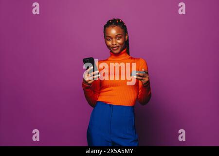 Jeune femme avec des tresses bicolores se tient sur un fond violet, en utilisant un smartphone pour effectuer un paiement en ligne sur une application de shopping. Banque D'Images