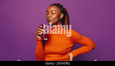 Femme heureuse se tient sur un fond violet, tenant une tasse avec une paille. Ses tresses bicolores tendance et son maquillage élégant ajoutent à son comportement joyeux. Banque D'Images