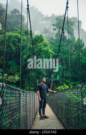 Un homme se tient à mi-chemin sur un pont suspendu brumeux entouré par la verdure luxuriante d'une forêt de nuages du Costa Rica, embrassant le surrou naturel tranquille Banque D'Images
