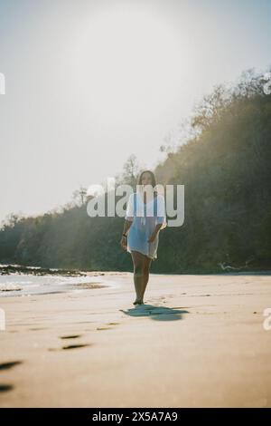 Une femme solitaire en blanc marche le long d'une plage déserte du Costa Rica, avec le soleil projetant une lueur chaude autour d'elle Banque D'Images