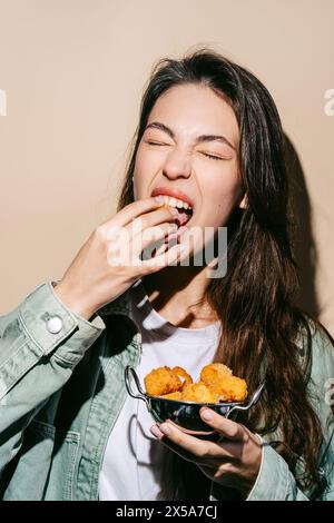 Une jeune femme joyeuse dans une tenue décontractée mordant dans une morsure de mozzarella frite dorée avec plaisir, tenant un bol de pièces supplémentaires. Banque D'Images