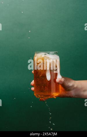 La main d'une femme recadrée méconnaissable tient une tasse en verre de bière légère avec de la mousse débordant sur un fond vert, capturant le mouvement dynamique O. Banque D'Images