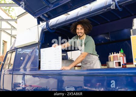 Un vendeur joyeux aux cheveux bouclés présente un menu dans un food truck bleu vibrant, incarnant la culture de la Street food à la mode Banque D'Images