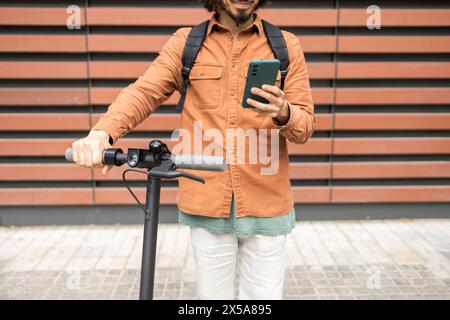 Un jeune homme en tenue décontractée interagit avec son smartphone tout en se tenant sur un scooter électrique, debout sur un fond géométrique Banque D'Images