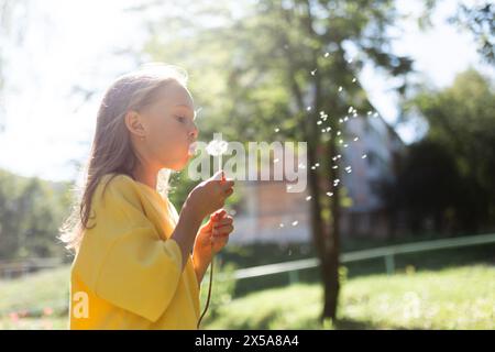 Une jeune fille dans un haut jaune souffle paisiblement un pissenlit dans un parc ensoleillé, avec des graines dispersées dans l'air Banque D'Images