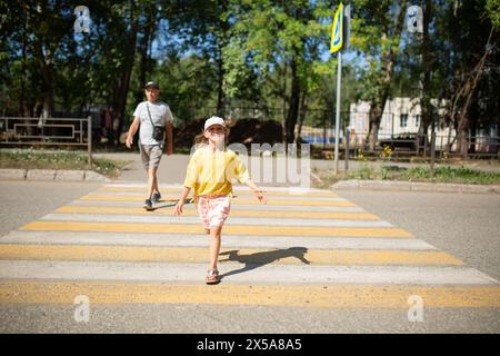 Une jeune fille joyeuse dans un haut jaune et une casquette saute joyeusement à travers une traversée de zèbre par une journée ensoleillée brillante, avec son grand-père marchant derrière elle Banque D'Images