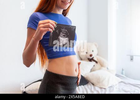 Jeune femme enceinte souriant tout en tenant une image échographique, avec un ours en peluche en arrière-plan Banque D'Images