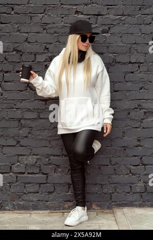 Une femme blonde cool en tenue décontractée profite de son café contre un mur de briques texturées. Banque D'Images
