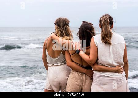 Vue arrière de trois femmes méconnaissables qui se tiennent à proximité, s'embrassant tout en regardant la mer, montrant l'amitié et le soutien. Banque D'Images