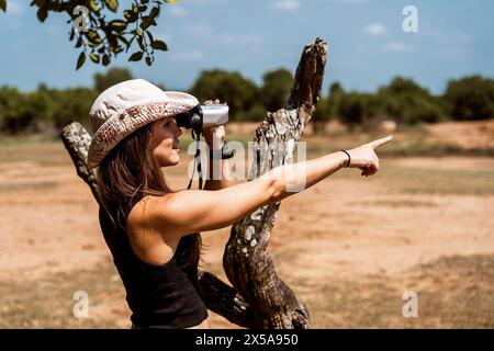 Femme dans un chapeau de safari utilise des jumelles pour observer la faune tout en pointant dans la distance, contre un paysage de brousse naturel. Banque D'Images