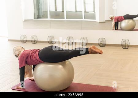 Vue de côté d'une femme musulmane concentrée dans un hijab s'engage dans une pose de yoga en utilisant une balle de stabilité dans un studio lumineux et moderne. Banque D'Images
