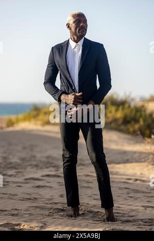 Un mannequin afro-américain pose en toute confiance sur une plage de sable, vêtu d'un élégant costume noir et d'une chemise blanche Banque D'Images