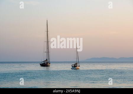 Deux voiliers flottent calmement sur la mer tranquille au large de la côte de Sardaigne, en Italie, tandis que le coucher de soleil peint le ciel dans des teintes douces Banque D'Images