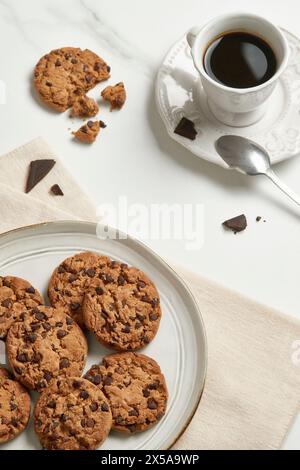 Un arrangement sophistiqué avec une tasse de café noir et des biscuits aux pépites de chocolat sur une assiette en céramique, avec des éclats de chocolat noir à proximité Banque D'Images