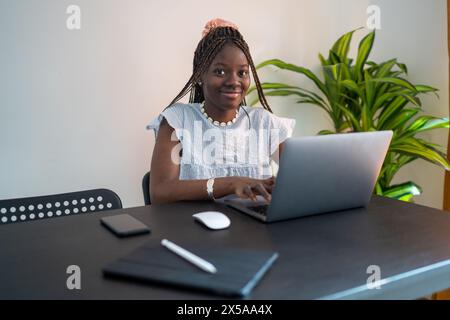 Une femme noire souriante avec des cheveux tressés travaille sur son ordinateur portable dans une configuration de bureau à domicile, exsudant productivité et confort Banque D'Images