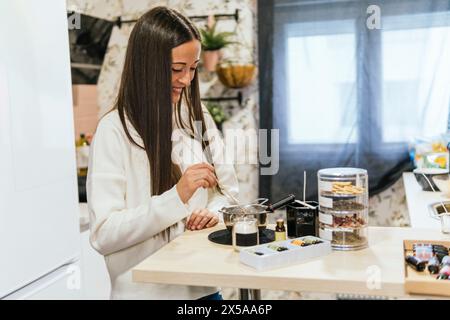 Une jeune femme joyeuse est montrée fabriquant des bougies maison dans un cadre de cuisine moderne et lumineux, rayonnant de positivité et de créativité Banque D'Images