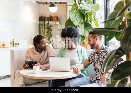 Trois jeunes professionnels s’engagent dans une discussion avec un ordinateur portable et une tablette numérique dans un espace de coworking bien éclairé entouré de verdure intérieure Banque D'Images
