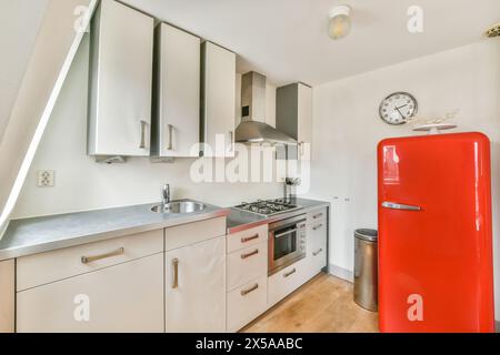 Comptoir avec évier et armoires blanches près du réfrigérateur rouge de style vintage dans la cuisine claire et élégante avec des appareils modernes et horloge dans l'appartement Banque D'Images