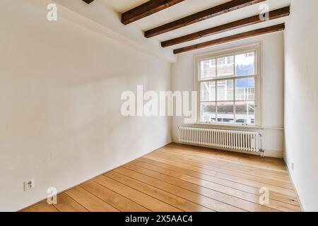 Cette chambre ouverte et ensoleillée présente du parquet ciré, des murs blancs et des poutres apparentes au plafond, une grande fenêtre et un radiateur. Banque D'Images