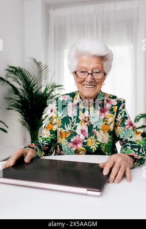 Une femme âgée joyeuse dans une chemise florale sourit alors qu'elle travaille sur un ordinateur portable dans un cadre lumineux à la maison, mettant en valeur la vie active des personnes âgées Banque D'Images
