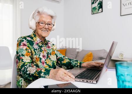 Joyeuse dame âgée avec des écouteurs utilise un ordinateur portable dans une pièce lumineuse et confortable, mettant en valeur le vieillissement actif et les personnes âgées technophiles Banque D'Images