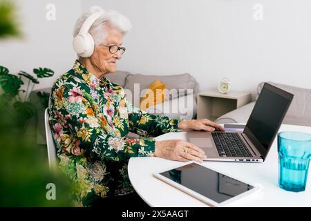 Une femme âgée travaille à la maison sur son ordinateur portable tout en portant des écouteurs, mettant en valeur l'utilisation de la technologie moderne par les aînés Banque D'Images