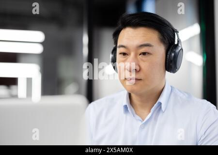 Un homme asiatique professionnel en tenue d'affaires se concentre intensément tout en portant des écouteurs. Il semble engagé et sérieux, assis dans un bureau moderne. Banque D'Images