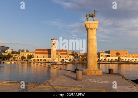 Rhodes, Grèce - 12 juin 2023 : entrée du port de Rhodes avec statue médiévale et maisons au soleil du matin. Banque D'Images