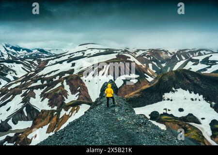 Réussi de randonneur féminin en veste jaune debout sur le sentier Blanhjukur avec montagne volcanique et neige couverte dans la journée sombre à Landmannalaugar, Icel Banque D'Images