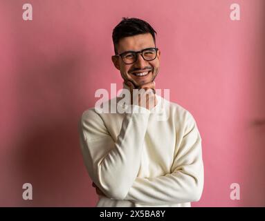 Un homme avec des lunettes et un pull blanc se tient devant un mur rose, souriant Banque D'Images