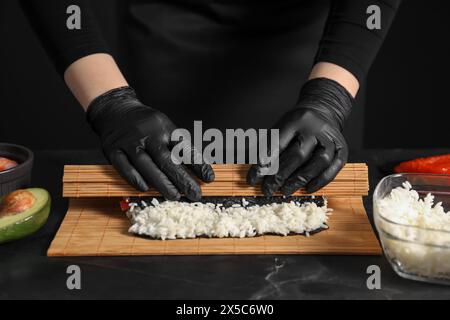 Chef en gants enveloppant le rouleau de sushi à la table texturée sombre, gros plan Banque D'Images