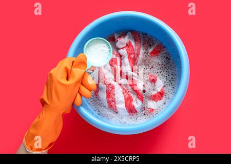 Femme dans des gants en caoutchouc ajoutant du détergent à lessive aux vêtements dans un bassin en plastique sur fond rouge Banque D'Images