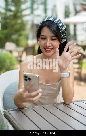 Une femme asiatique attrayante dans une robe mignonne agite la main, saluant quelqu'un par un appel vidéo sur son smartphone, parlant pendant un appel vidéo pendant que R Banque D'Images