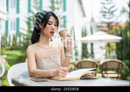 Une jolie femme asiatique dans une robe mignonne écrit dans son journal et apprécie le café tout en étant assise à une table dans un beau jardin. loisirs et style de vie Banque D'Images