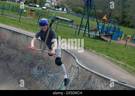 Jeune en chemise verte et casque bleu monte son scooter dans les airs au Wilderness Skate Park, Porthcawl, Royaume-Uni. 27 avril 2024. Banque D'Images