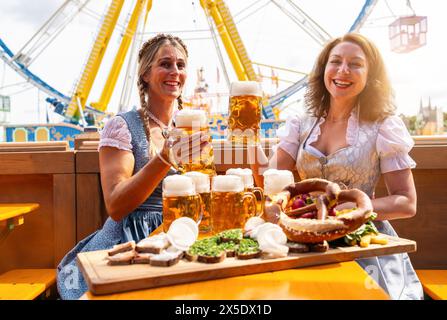 Deux femmes en robes bavaroises traditionnelles ou tracht toasting avec des tasses à bière et de la bière et des collations sur la table à un parc d'expositions festif en Bavière oktob Banque D'Images