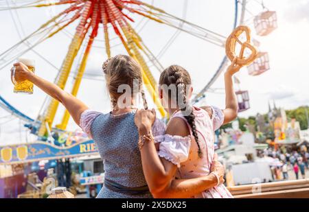 Des femmes bavaroises à tracht tenant une tasse de bière et un bretzel en altitude dans un champ de foire animé avec grande roue à l'oktoberfest ou au duld en allemagne Banque D'Images